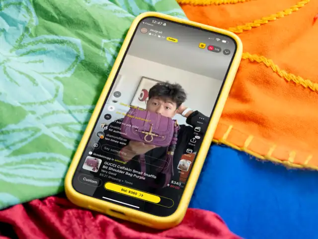 A smartphone with a yellow case displays a livestream shopping event on a popular shopping app, featuring a person presenting a purple handbag placed on colorful fabric.